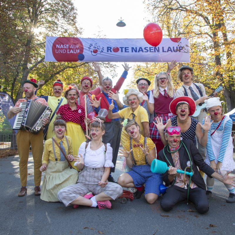 Eine Gruppe ROTE NASEN Clowns posiert vor dem Start Banner des ROTE NASEN Laufs.